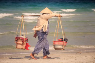 yan görünümü Yerel satıcı kadın boyunduruğu potalara ile şapkalı masmavi deniz dalga sörf karşı kum plaj devam ediyor