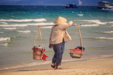 masmavi deniz tekne yüzen ev karşı plaj boyunca closeup Vietnamca satıcı kadın boyunduruğu kova ile şapkalı yürür
