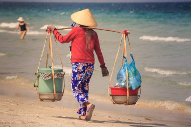 Deniz karşı plaj boyunca closeup arka görünümü yerel meyve satıcı kadın boyunduruğu kovalar ile ulusal beyaz şapkalı yürür