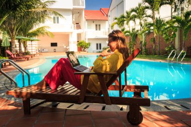 otel yüzme havuzunda sandalyenin altına gölge dizüstü bilgisayar ile katlama elbiseleri sarı uzun saçlı kız oturur