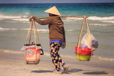 masmavi deniz karşı kum plaj boyunca closeup arka Vietnamca meyve satıcı kadın boyunduruğu kova ile şapkalı yürür