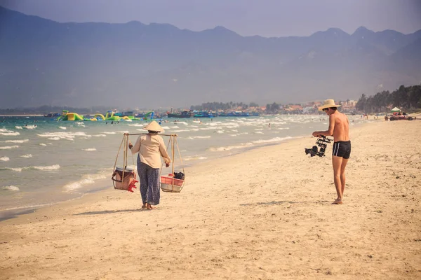 European young man photos with professional camera Vietnamese vendor woman with yoke on beach against wave crests