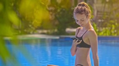 telefon gülümsüyor Yüzme otel havuzu ve bitkiler karşı güneş ışınları altında at kuyruğu görüşmeler ile portre sarışın kız