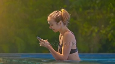 güzel kız havuzu güneş altında internet sörf için smartphone kullanır closeup yan görünüm karşı ağaçlar ışınları