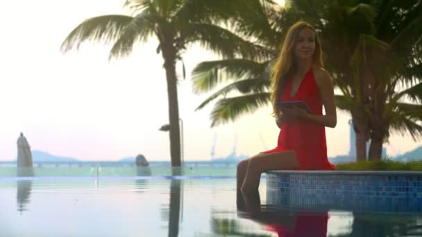年轻漂亮的金发女郎穿着红色礼服坐在酒店泳池边缘在阳光下的棕榈和大海 — 图库视频影像