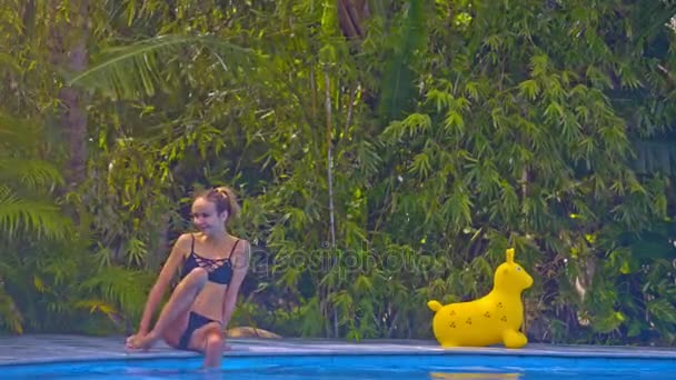 美丽的女孩在黑色泳装坐在水池边缘和倾斜的腿入水反对绿色植物在晴朗的天 — 图库视频影像