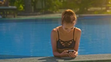 güzel gülümseyen ince sarışın kız bikini ile smartphone mavi su yüzme havuzu parapetin üzerinde sıcak güneşli günde eğilir.