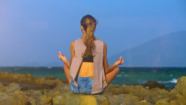 背面视图苗条的女孩长马尾辫坐在莲花姿势在岩石海滩看图案蔚蓝无边的海洋 — 图库视频影像