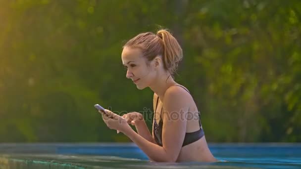 侧面视图美丽的微笑的女孩看着智能手机和休息在 Waterpool 的绿色树木 — 图库视频影像