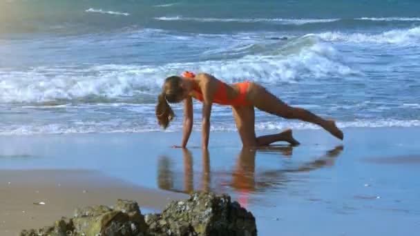 うねる泡波に対しての濡れた砂浜に膝の上のオレンジ色の水着列車でポニーテールのスリムな金髪女の子 — ストック動画