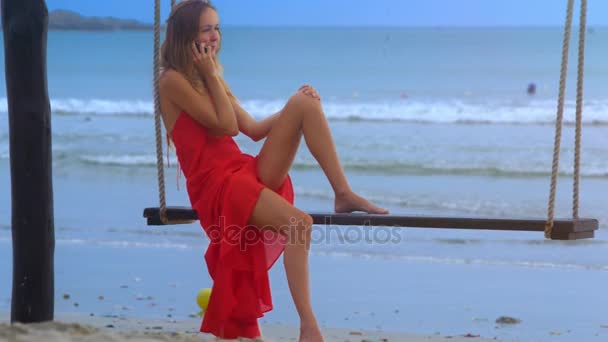 特写侧面视图苗条的女孩在透明的红色裙摇摆在海滩长凳讲话在电话反对泡沫波浪冲浪 — 图库视频影像