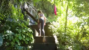 güneş ışığına karşı vahşi tropik bitkiler arasında Ahşap merdiven için yan görünümü ince sarışın kız uzun at kuyruğu ile çıkageldi