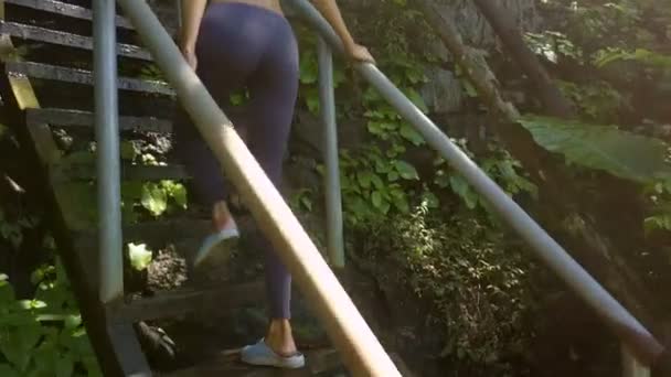 底部视图苗条的长腿女孩爬上古老的木制楼梯 用栏杆在热带植物中对抗阳光 — 图库视频影像