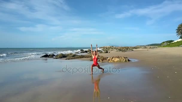 穿红色泳装的漂亮女孩练习瑜伽体式在海上潮湿的沙滩上 与图案海景和沙滩棕榈 — 图库视频影像