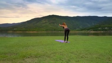 Flycam side görünümü genç ince sarışın kız yoga poz Utkatasana düz yeşil göl kıyısında tropikal doğaya karşı tutar