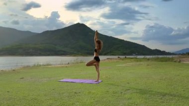 Flycam bir kenara yoga poz Vrikshasana parlak gri gökyüzü altında yeşil çim çayır üzerinde kız at kuyruğu ile gösterir