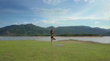 Flycam görünümü uzun boylu kız standları Yoga barışçı nehir güneş ışınları altında parlayan karşı Vrikshasana poz