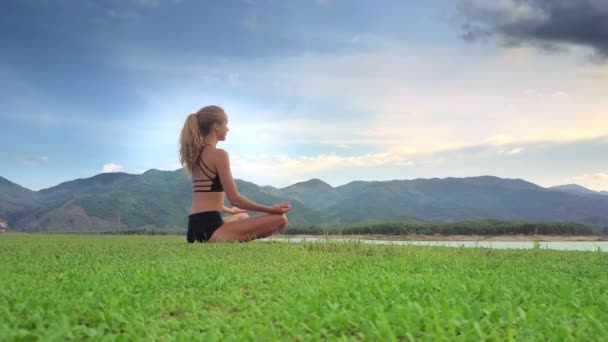 Flycam 背面视图年轻女孩举行瑜伽位置 Ardhapadmasana 湖岸反对惊人的丘陵景观 — 图库视频影像