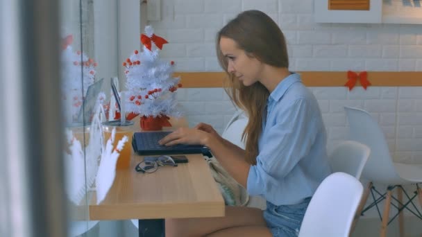 側面ビューかわいい女の子は青いシャツとデニム短パン小さなクリスマス ツリーに対して木製カウンターでコンピューター上の種類 — ストック動画