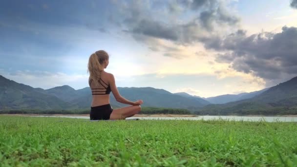 曇り空の下で湖銀行草原ヨガ ポーズ ロータス瞑想にポニーテールの裏面表示金髪少女 — ストック動画