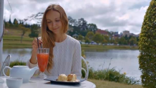 クローズ アップ笑みを浮かべて少女は 曇り空の下で緑の植物に対してカフェでスプーンで冷たいオレンジ飲料をかき立てる — ストック動画