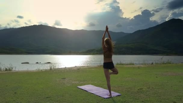 背面看苗条的金发女孩站在瑜伽姿势 Vrikshasana 在阳光湖下闪烁的垫子附近 — 图库视频影像