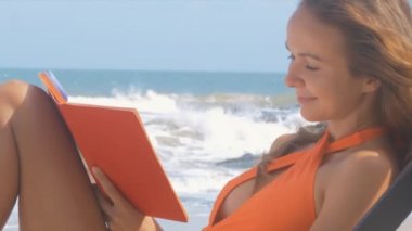 gülümsemek gülümseyen uzun saçlı kadın bikini defter görünüyor ve güneşli okyanus kıyısında yazıyor