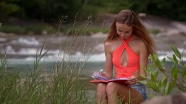 güzel sarışın uzun saçlı kız portakal Top Kağıt defter köpüklü nehir karşı diz ile rüzgarlı gün oturur