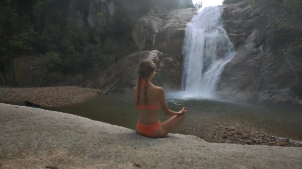 背面视图金发女郎长辫子比基尼坐在瑜伽姿势莲花在石头上反对瀑布 — 图库视频影像