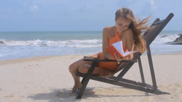 絵画画像強風揺れる彼女は海の泡波がビーチに折りたたみ椅子の日記と座っている女の子ブロンド髪 — ストック動画