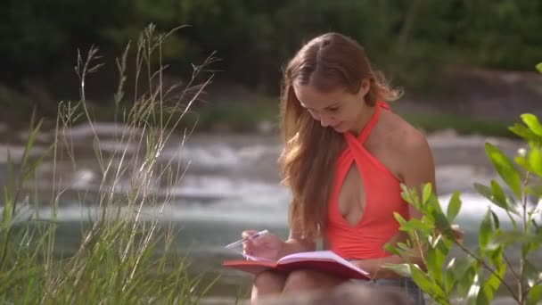 可爱的金发长发女孩在打开橙色顶部在河岸上的绿灌木丛中写笔记 — 图库视频影像
