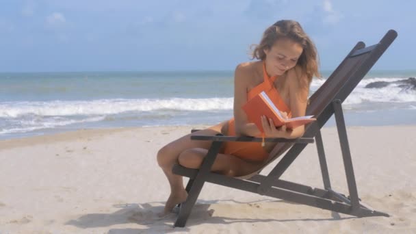 Blondes Mädchen mit langen Haaren sitzt im Liegestuhl und schreibt in Tagebuch am Sandstrand vor blauem Meer und klarem Himmel