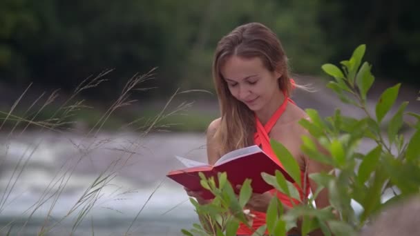 Atractiva Chica Rubia Concentrada Lee Libro Rojo Río Ramas Verdes — Vídeo de stock