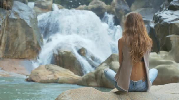 カスケードの滝の近くの石の上に座っているジーンズの長いゆったり流れる金髪の裏面表示女の子 — ストック動画