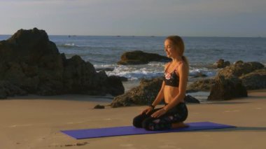yan görünümü güzel sarışın gülümseyen kız büyük kayalar ve okyanus karşı gergin yoga sonra mor mindere rahatlatır