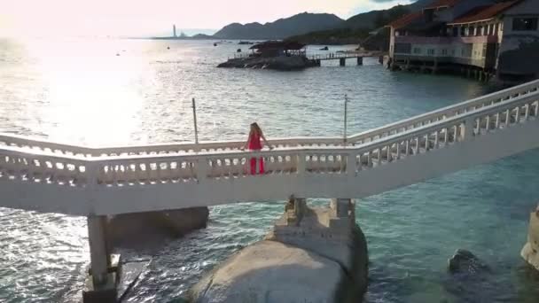 夕焼けと山に対する洋上白い橋の上長い赤いドレスでパノラマ ビュー女性が立っています — ストック動画