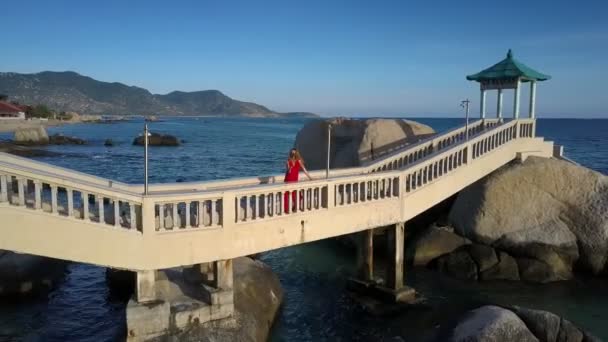 赤のディナー ドレスの上部表示金髪長い髪の女の子ポーズに対して無限のターコイズ ブルーの海の石の橋 — ストック動画