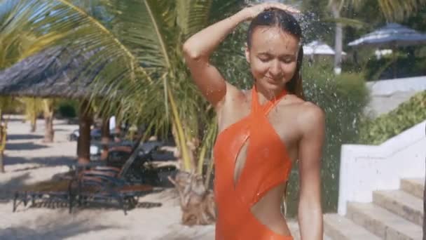 オレンジ色の水着で魅力的な女の子が砂のビーチ パラソルとヤシの木のシャワー ストリームの下に立つ — ストック動画