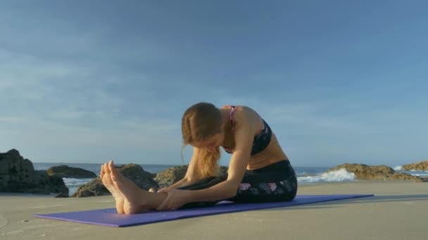 美しいスリムなブロンドの女の子はマットの上に座っているし 砂のビーチで青空の石の間で膝に体を曲げる — ストック動画
