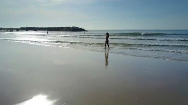 Mayo ince kadın okyanus ıslak kum plaj boyunca yürür ve parlak parlayan güneş ışınlarına karşı uzun saç düzeltir
