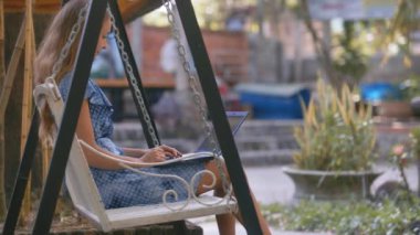 yan görünümü uzun saçlı kız mavi elbiseli beyaz salıncağa oturur ve tropikal bitki karşı modern dizüstü kapatır