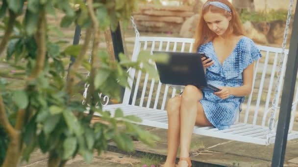 蓝色衣服上的金发女郎手持现代膝上型笔记本电脑在膝上的白色秋千上坐在绿色的树前 — 图库视频影像
