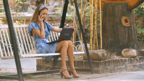 青いブランコ ベンチで緩い流れる髪の少女ノート パソコンを閉じ 木の彫刻公園で近く電話で話す — ストック動画