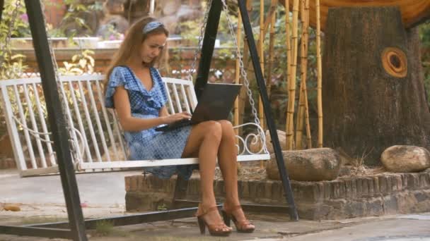 素敵な白いベンチ スイングと木の幹の近くのラップトップ型に座っている青いドレスのかなりブロンドの長い髪の女の子 — ストック動画