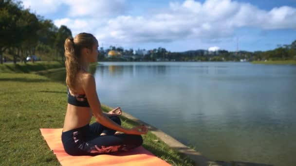 特写运动女孩坐在靠近湖的瑜伽位置 天空反射对遥远的城市景观 — 图库视频影像