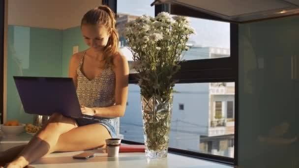 快乐的年轻女士看着白色的菊花和工作在笔记本电脑对窗口厨房 — 图库视频影像
