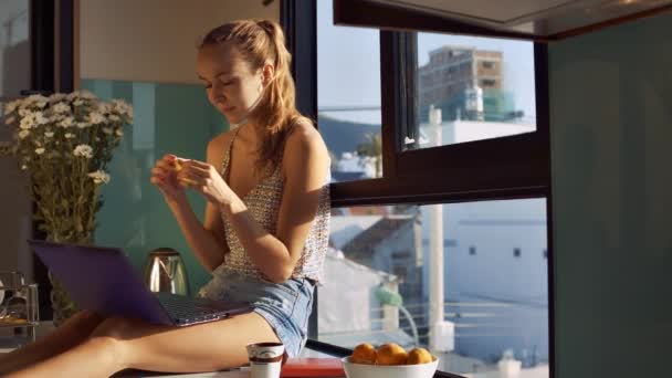 年轻快乐妇女在短裤吃香蕉并且存放笔记本电脑在窗台反对城市外面玻璃 — 图库视频影像