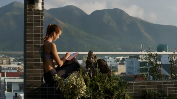 长辫子的金发女人倾斜在屋顶露台专栏和写在日志反对美妙的山在城市后面 — 图库视频影像
