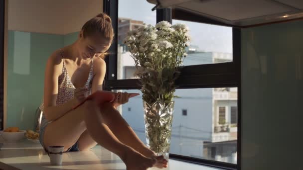 白い花とクリスタルの花瓶に近い日記窓辺にきれいな脚でかなりブロンドの女の子が裸足に座ってください — ストック動画