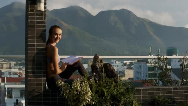Young blonde woman reads book sitting on roof terrace looks at camera man against pictorial hills and city — Stock Footage Young Blonde Woman Reads Book Sitting Roof Terrace Looks Camera Stock Footage